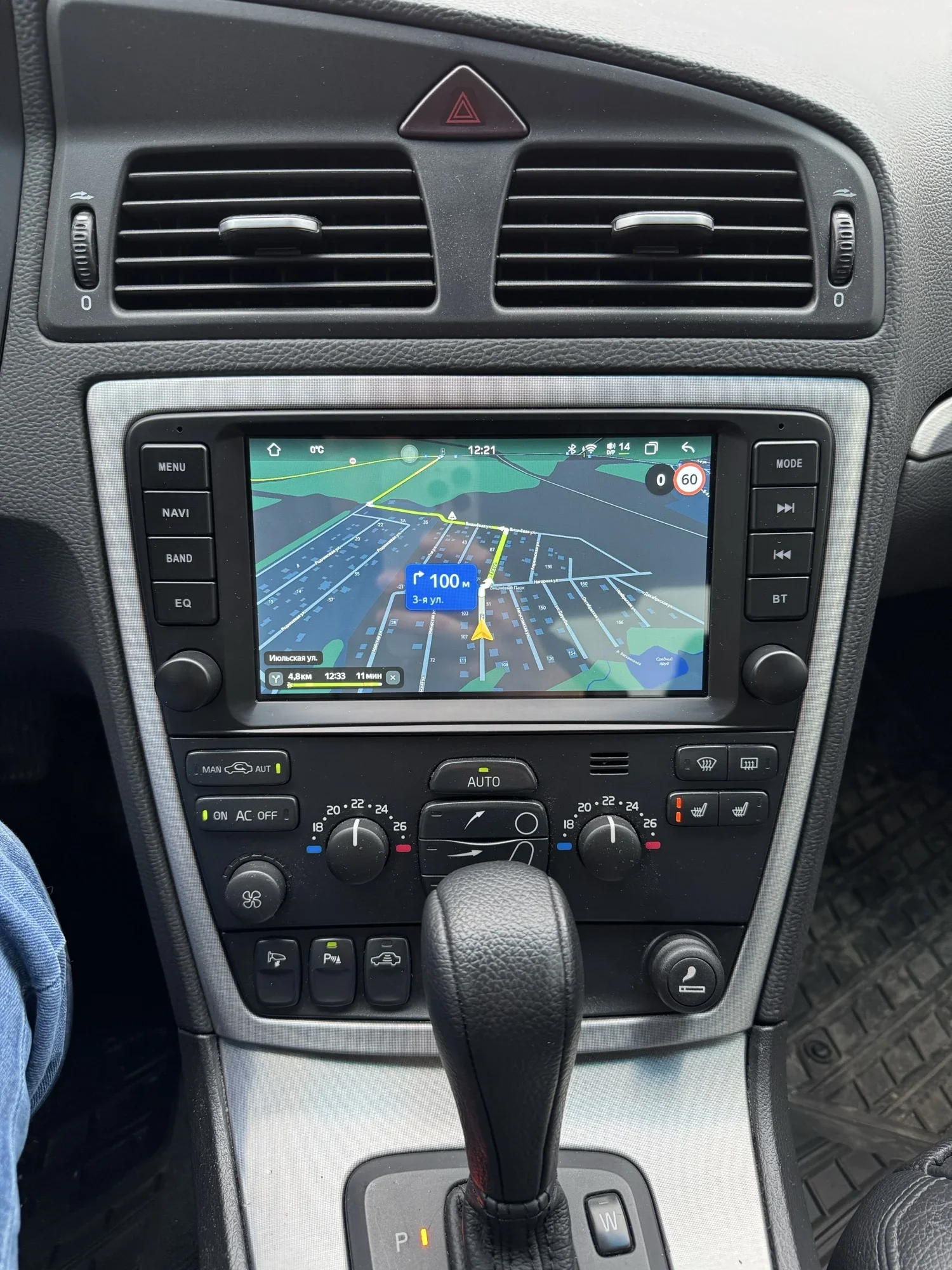 Car infotainment screen displaying a 3D navigation map with a blue '100 m' marker and upcoming turn, surrounded by climate controls and vents in a car interior.