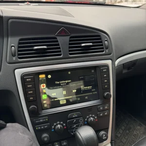 Car interior focusing on the touchscreen showing music, with climate controls and gear shifter in the foreground and a Christmas tree outside the windshield.