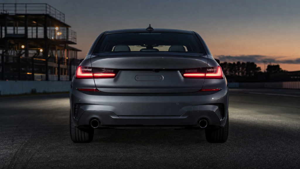 Rear view of a silver sedan parked on a track at dusk, with glowing red taillights and a dark sky in the background