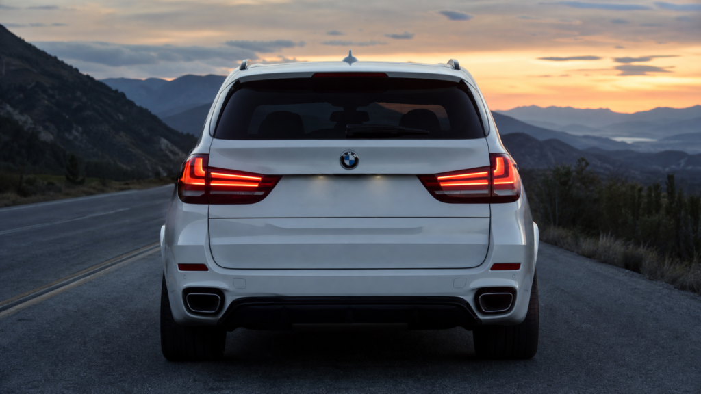 Rear view of a white BMW SUV on a mountain road at sunset, taillights illuminated, distant mountain ranges visible.