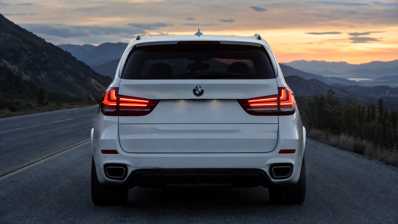 led-tail-lights-bmw-x5-f15-guide - speedXperts Rear view of a white BMW SUV on a mountain road at sunset, taillights illuminated, distant mountain ranges visible.